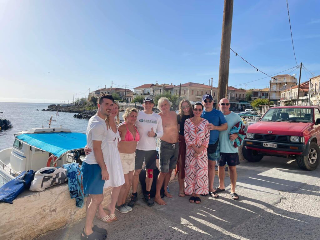 Group of smiling people posing together after a snorkeling and fishing trip in Kardamili, with a boat in the background and the quaint coastal town visible on a sunny day.