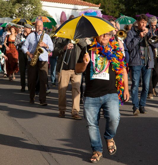 Man leading a colorful street parade with an umbrella and feather boa, followed by a marching jazz band.