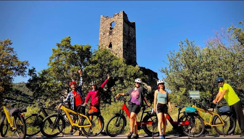 Group of cyclists with bikes posing in front of a historic stone tower during a cycling tour with 2407M in Kardamili