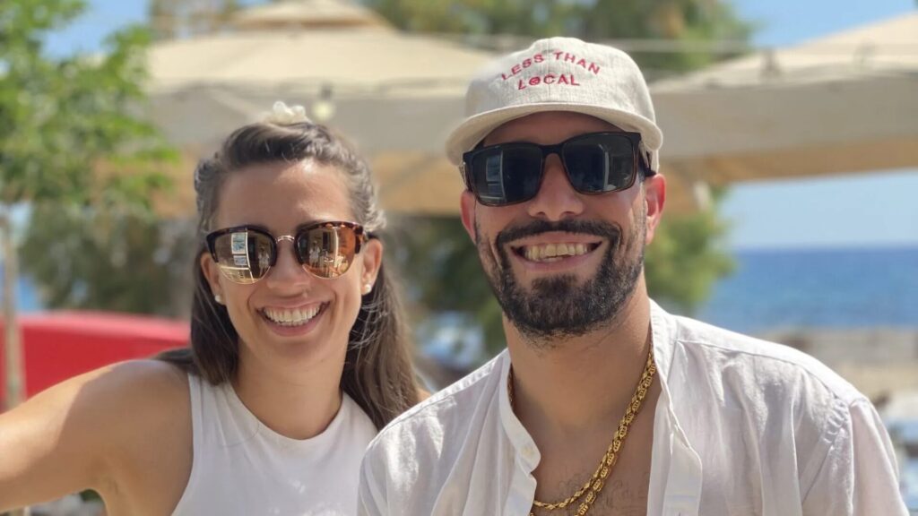 A happy couple enjoying their summer vacation in Kardamili, Greece, standing under the bright sun near the coast. The backdrop features a clear blue sky and hints of the Mediterranean Sea, radiating summer vibes.