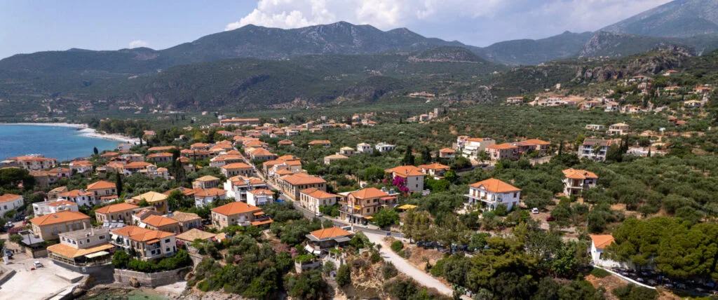 Aerial view of Kardamili village showcasing its red-tiled roofs, olive groves, and crystal-clear waters along the coastline, set against a backdrop of mountainous terrain.