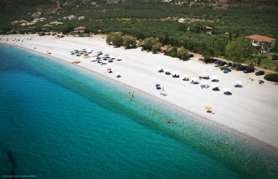 Aerial view of Ritsa Beach in Kardamili, Greece, with turquoise water, white pebbles, and scattered umbrellas