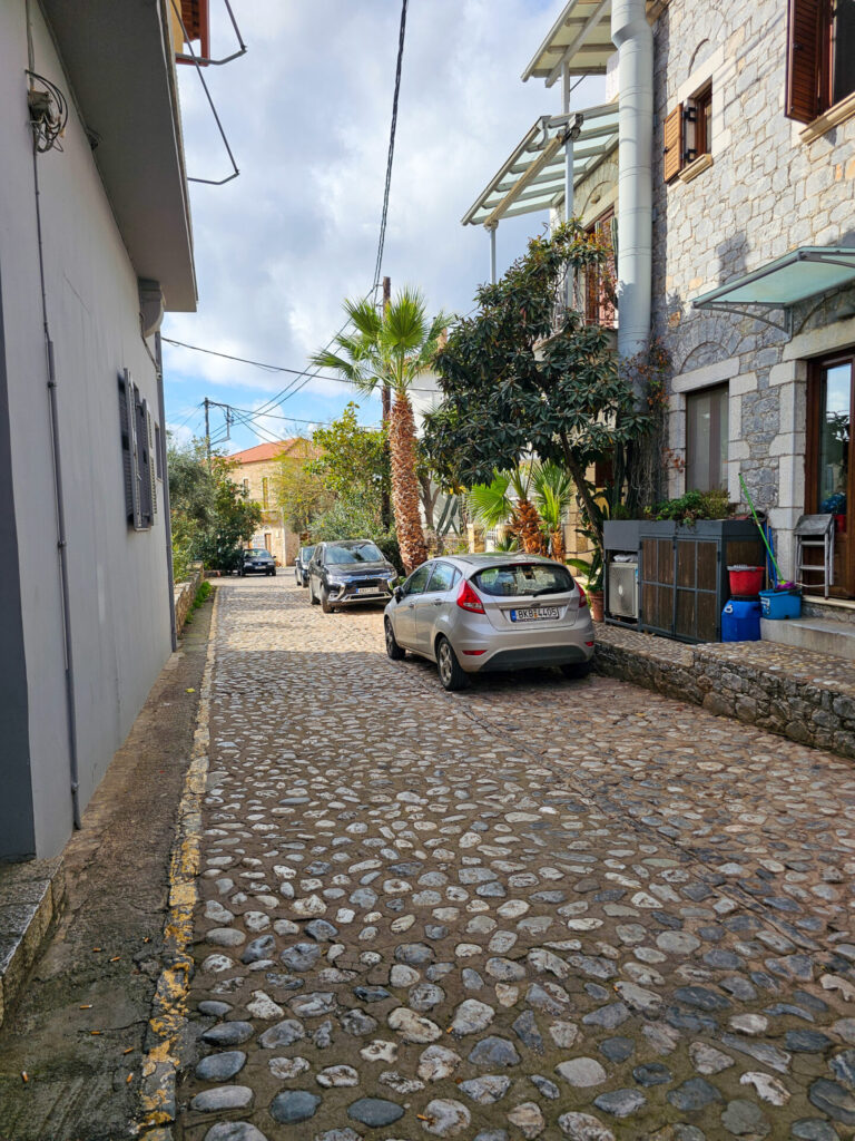 A narrow stone alleyway in Old Town Kardamili, lined with historic stone buildings, potted plants, and soft afternoon light under a blue sky.