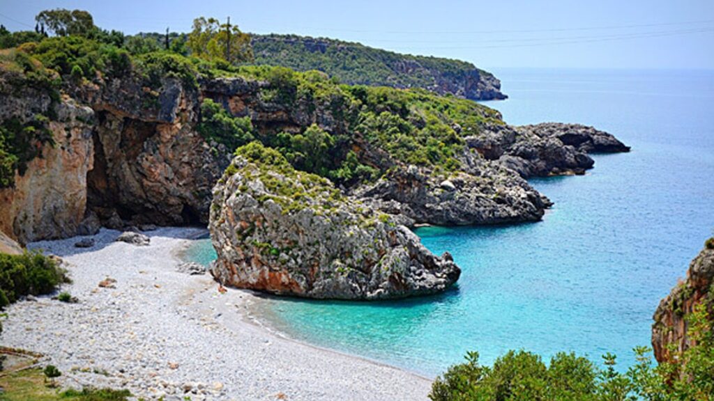 Foneas Beach in Kardamili with its iconic rocky outcrop, clear turquoise waters, and dramatic coastal cliffs on a sunny day.