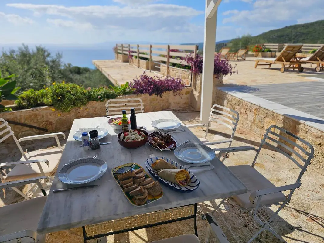 A luxury villa terrace featuring a high-end outdoor dining table overlooking the panoramic blue waters of the Messinian Gulf.