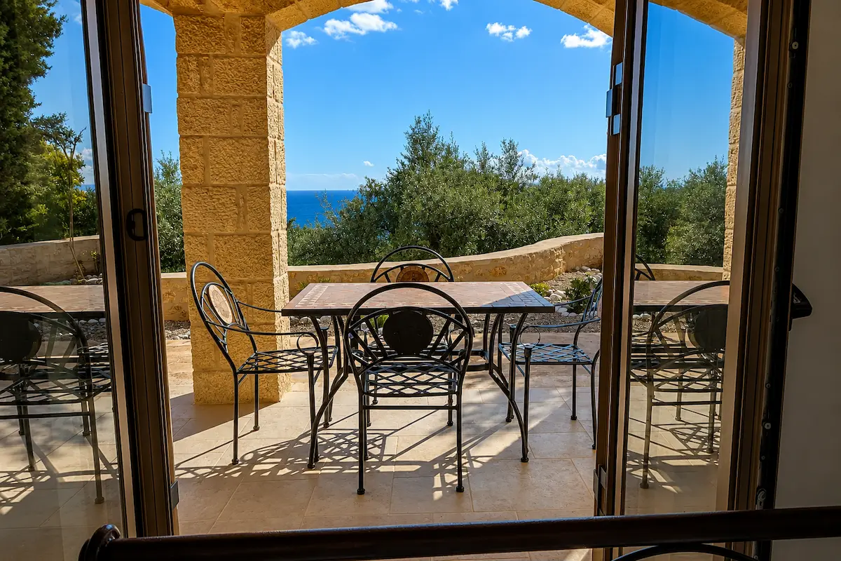 View from a Kardamyli villa overlooking a sunny stone terrace with outdoor dining furniture, olive trees, and the Mediterranean Sea in Mani, Greece.