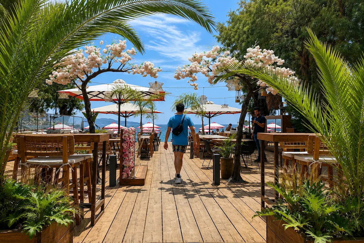 Sunny seaside café in Kardamyli in May with wooden decking, tropical plants, flowering arches, beach umbrellas, and visitors enjoying the Mediterranean coast under a blue sky.